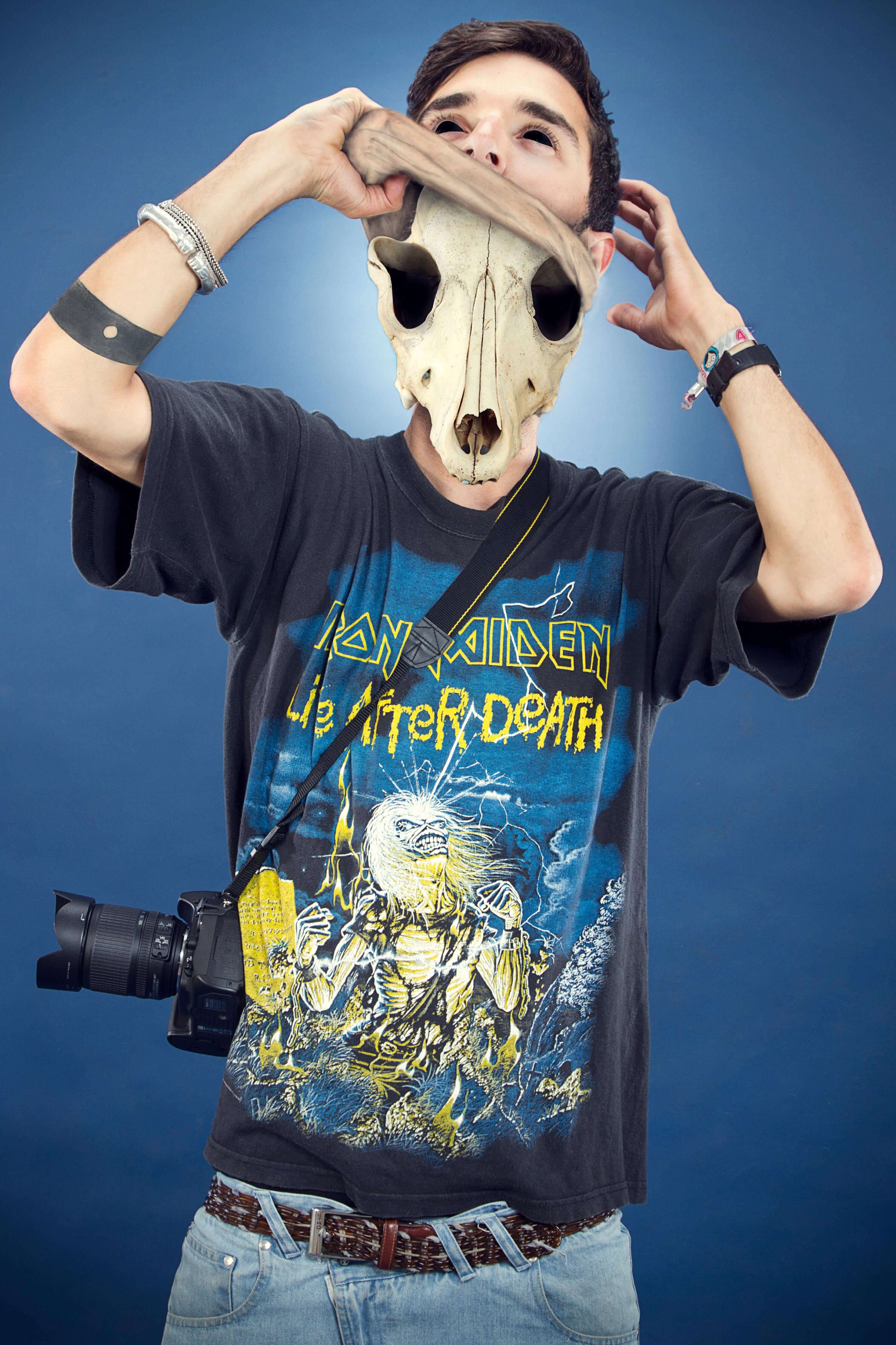 The man lifting the animal skull from his face, wearing a vintage band t-shirt with a camera slung across his chest, against a blue backdrop