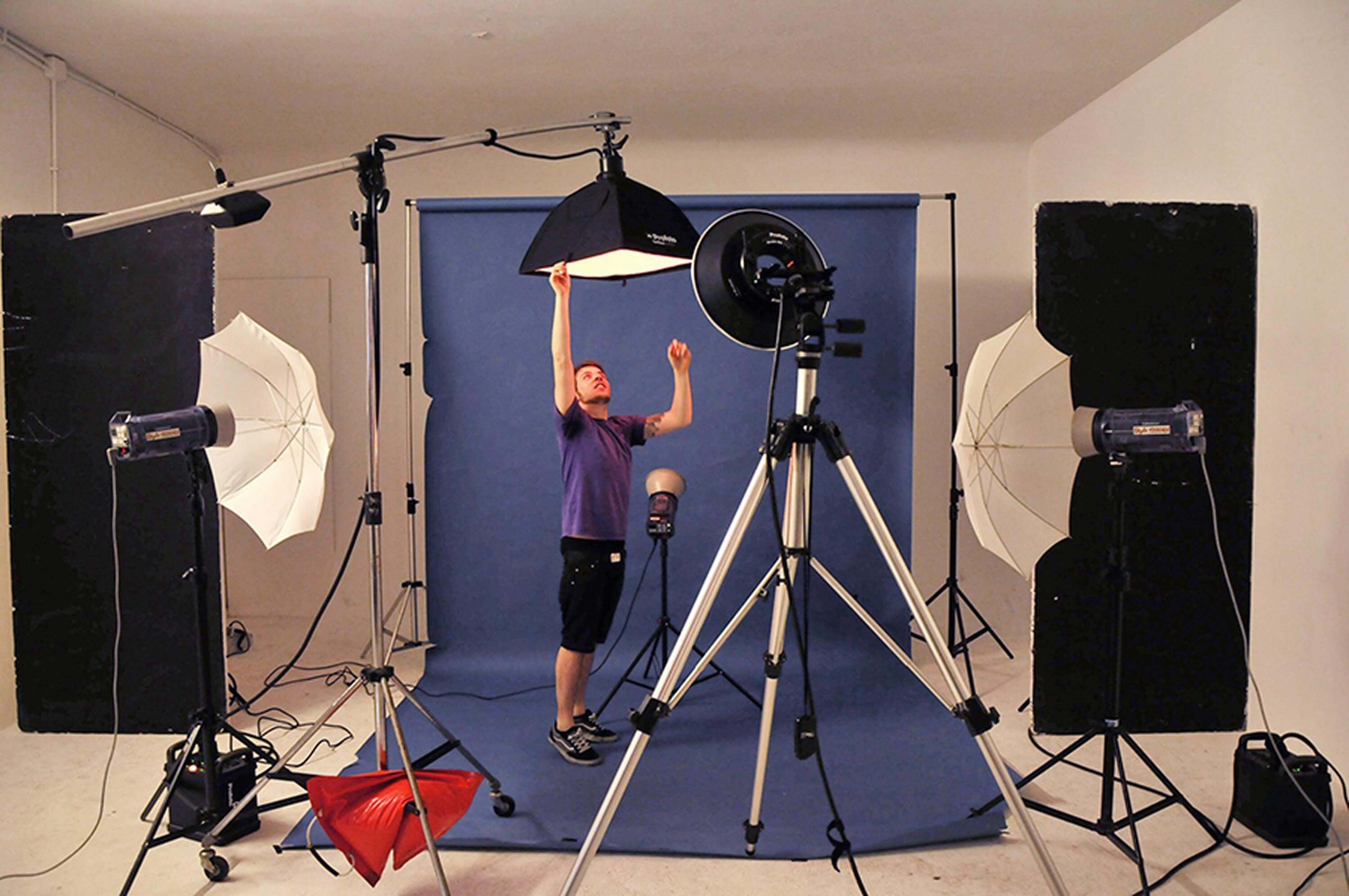 Behind the scenes — the photographer adjusting a boom-mounted softbox in a studio with a blue backdrop, umbrellas and lights around