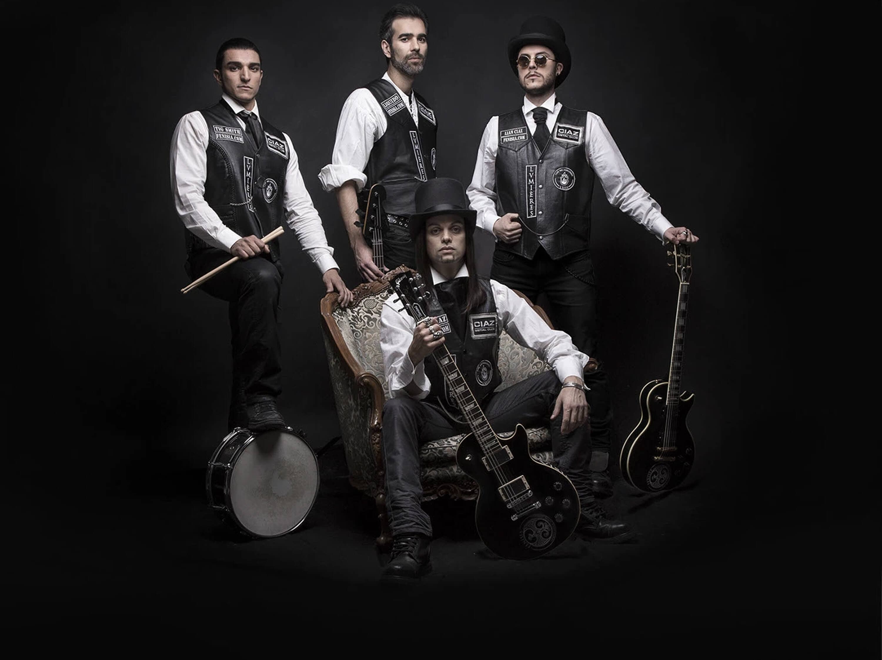 Studio group portrait — the four musicians with drumsticks, snare drum and two electric guitars, one seated on a brocade armchair, all in top hats and patched leather waistcoats