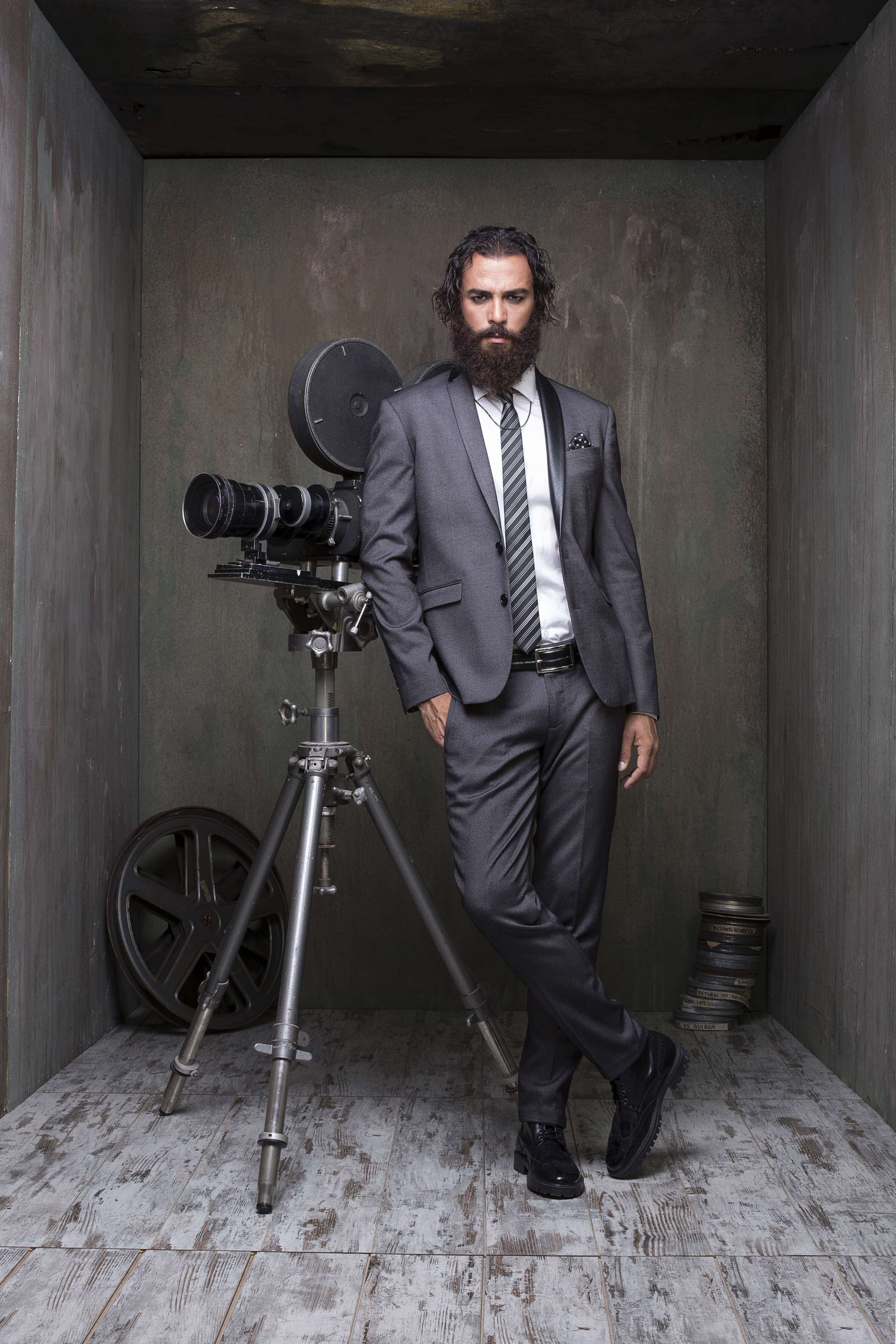 Grey suit and striped tie, the model leaning on a vintage film camera on a tripod with film reels, in a weathered concrete room