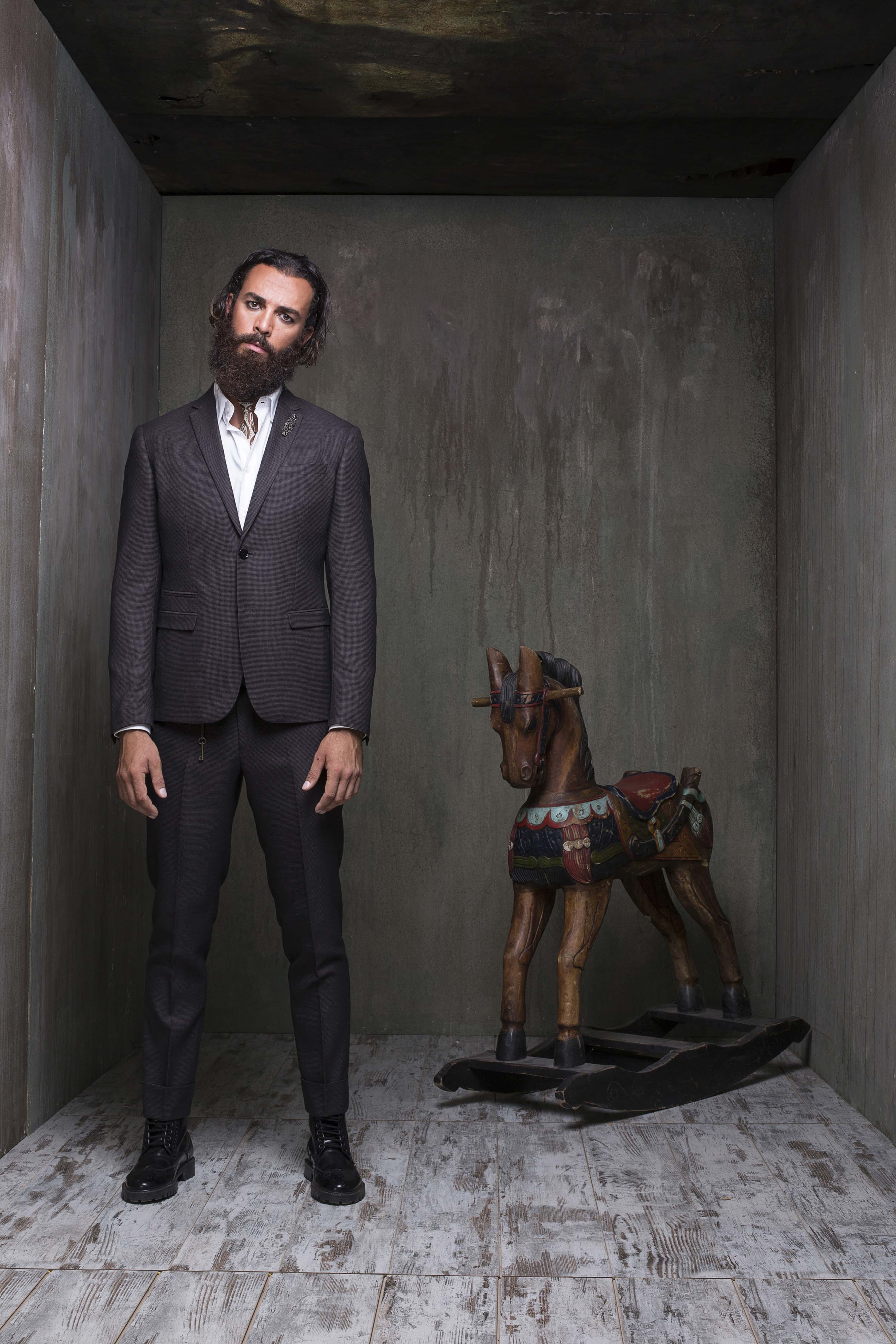 Charcoal suit standing beside a carved wooden rocking horse in the concrete room