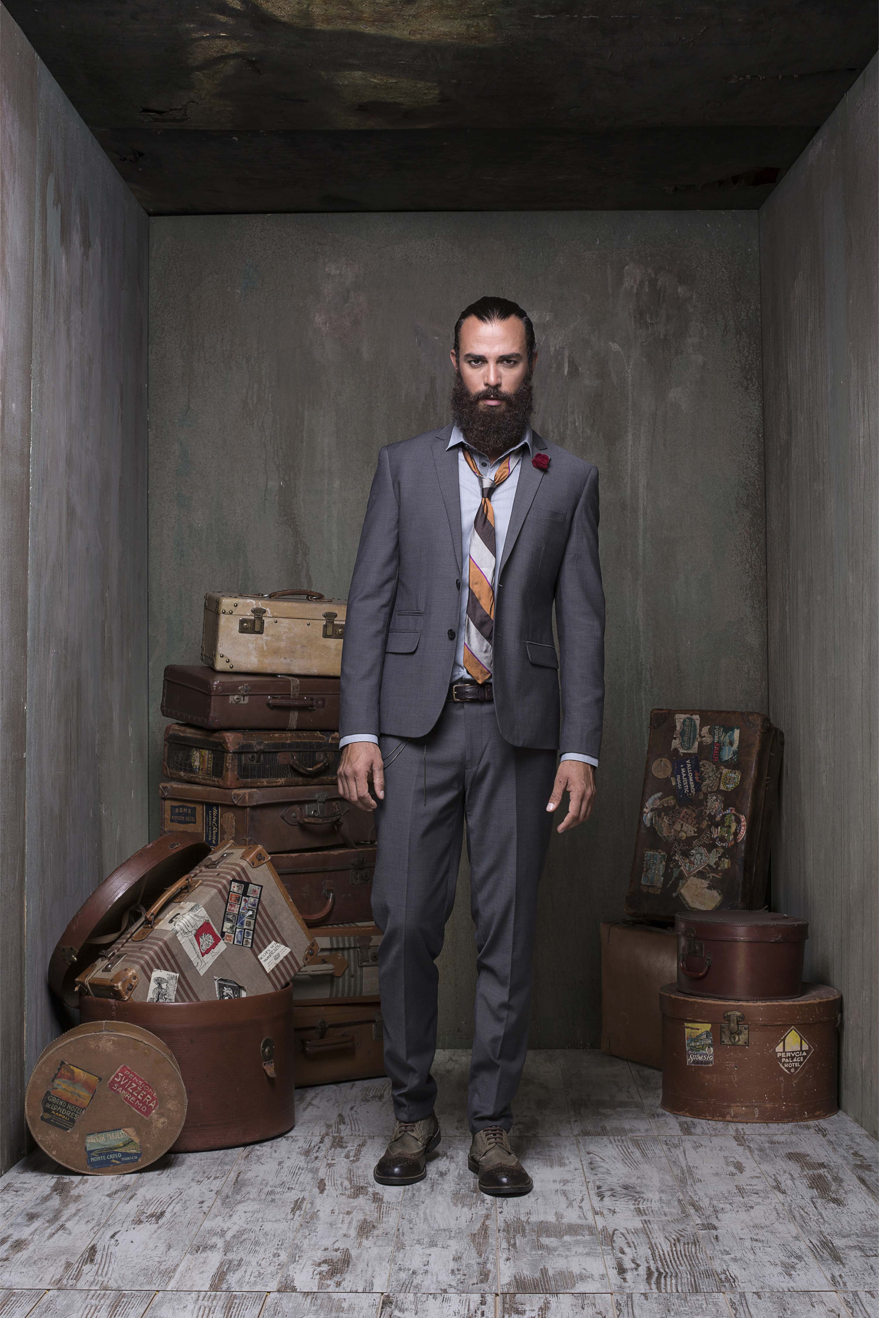 Grey suit with a striped tie and a red boutonniere, beside a stack of vintage suitcases and hat boxes