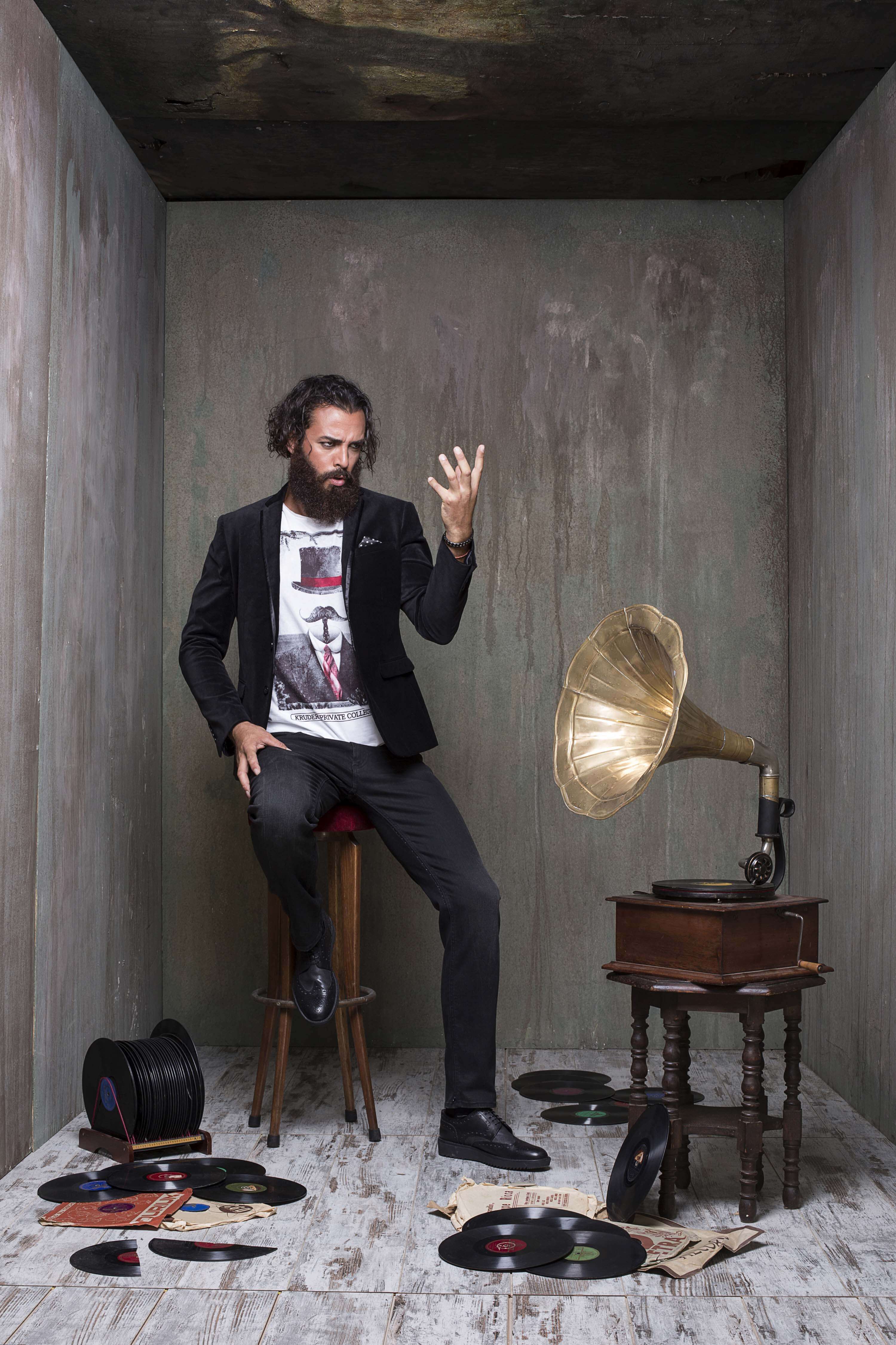 Black blazer over a printed tee, seated on a stool beside a brass horn gramophone and scattered vinyl records