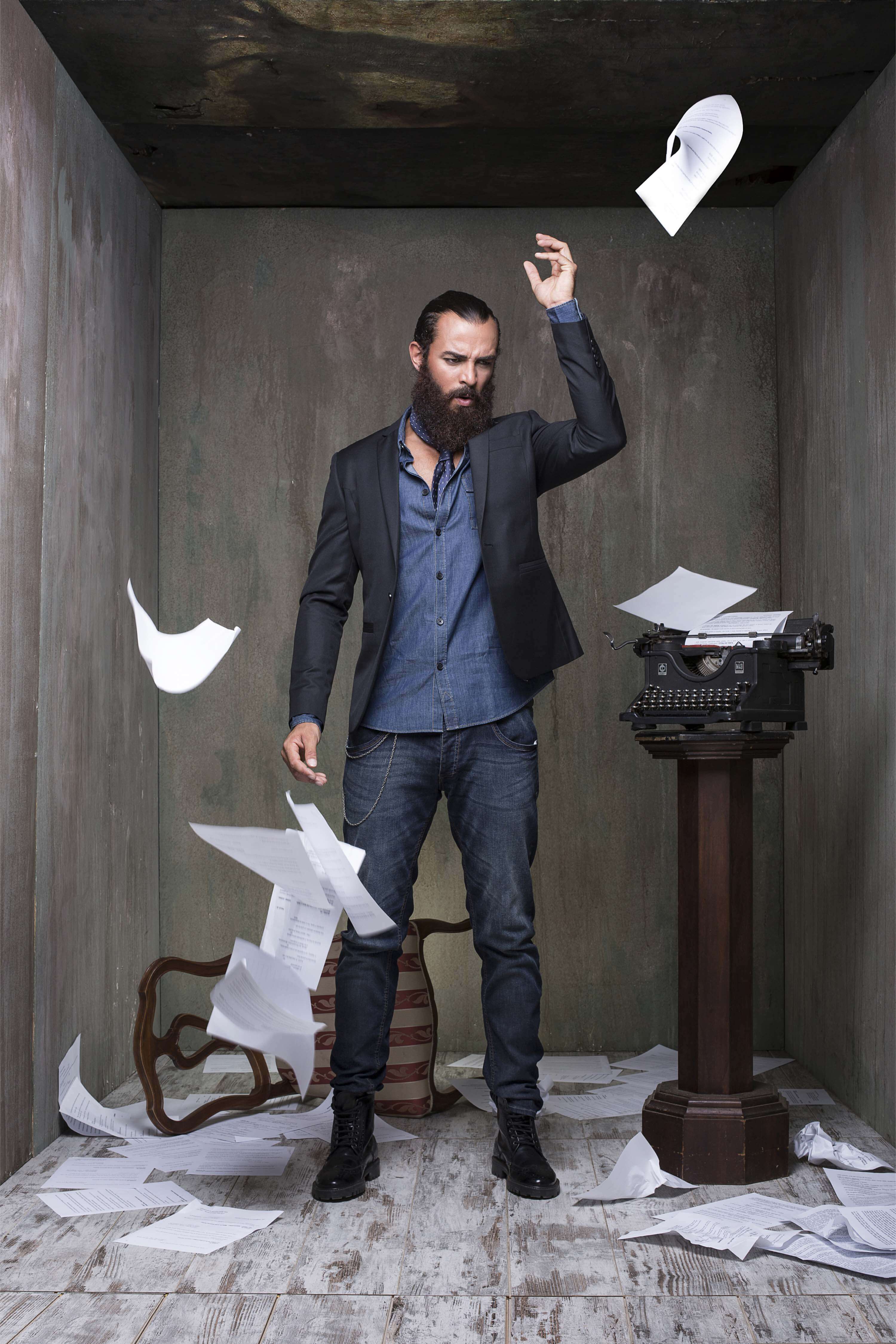 Dark blazer over a denim shirt and jeans, sheets of paper flying around him beside an old typewriter on a plinth