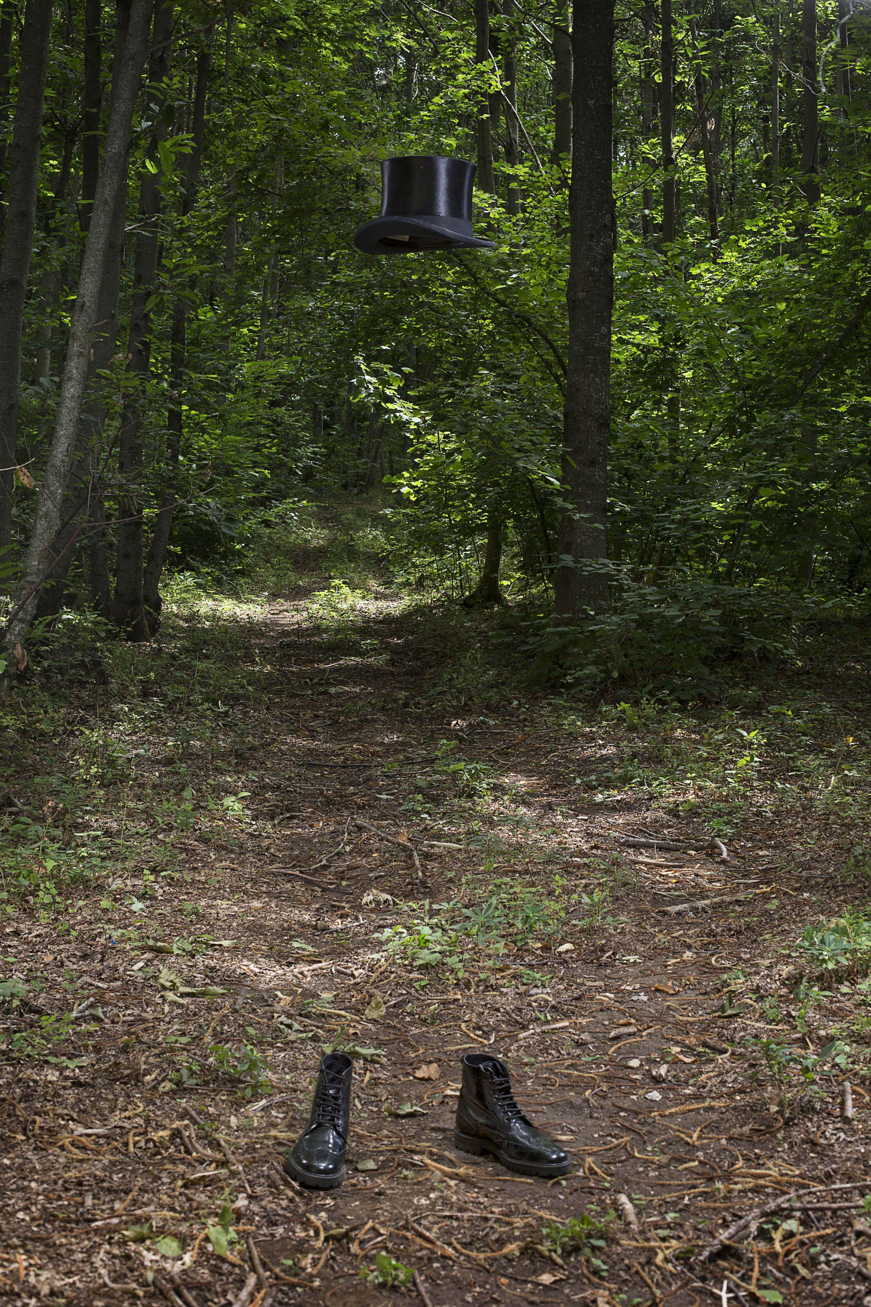 An empty forest path with a black top hat floating in the air above a pair of laced boots