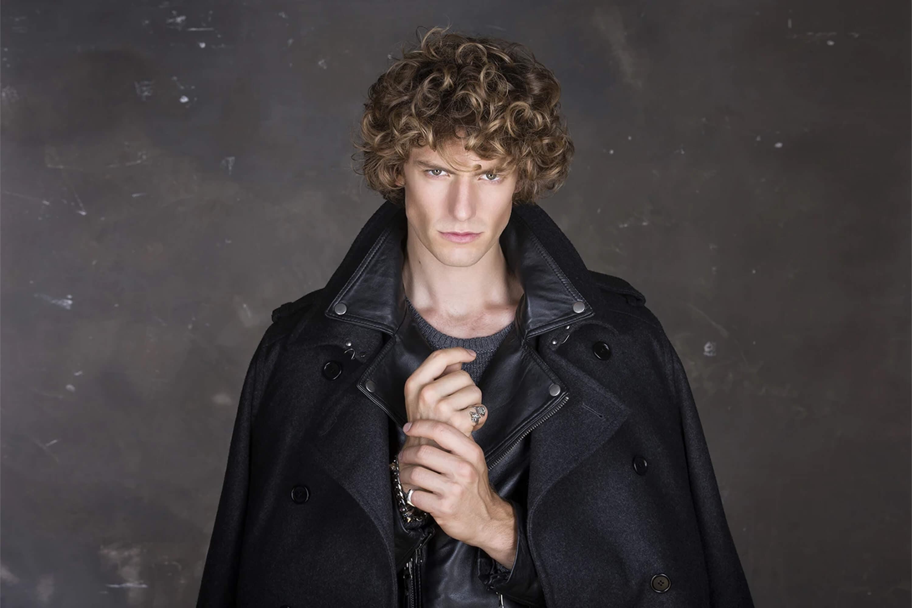 The model in a dark wool overcoat over a leather biker jacket and grey knit, hands raised to the collar with a signet ring, against the grey concrete wall