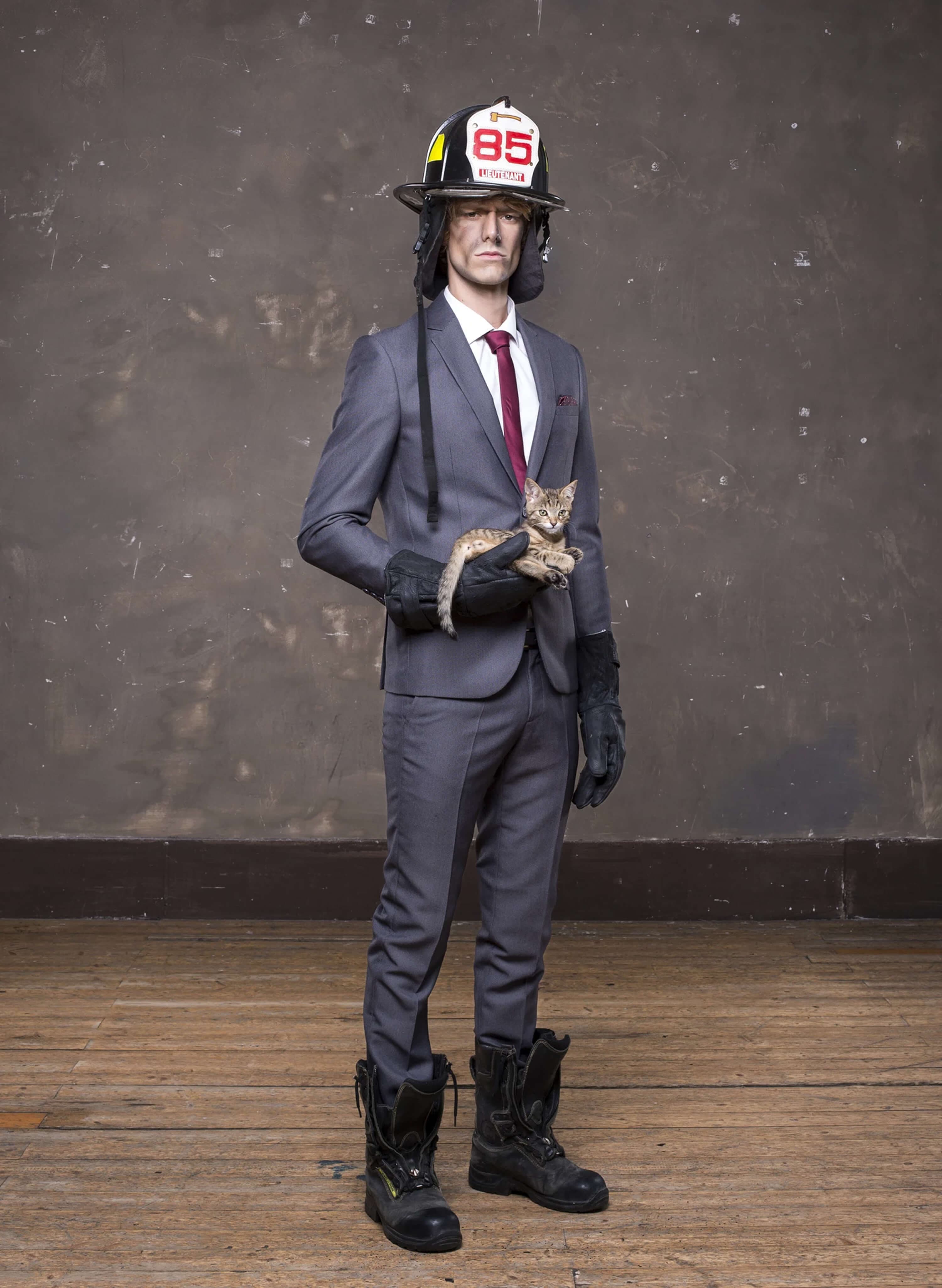 Full-length shot — grey suit with burgundy tie and firefighter helmet, black gloves and heavy fire boots, holding the kitten, on the worn wooden floor of the studio