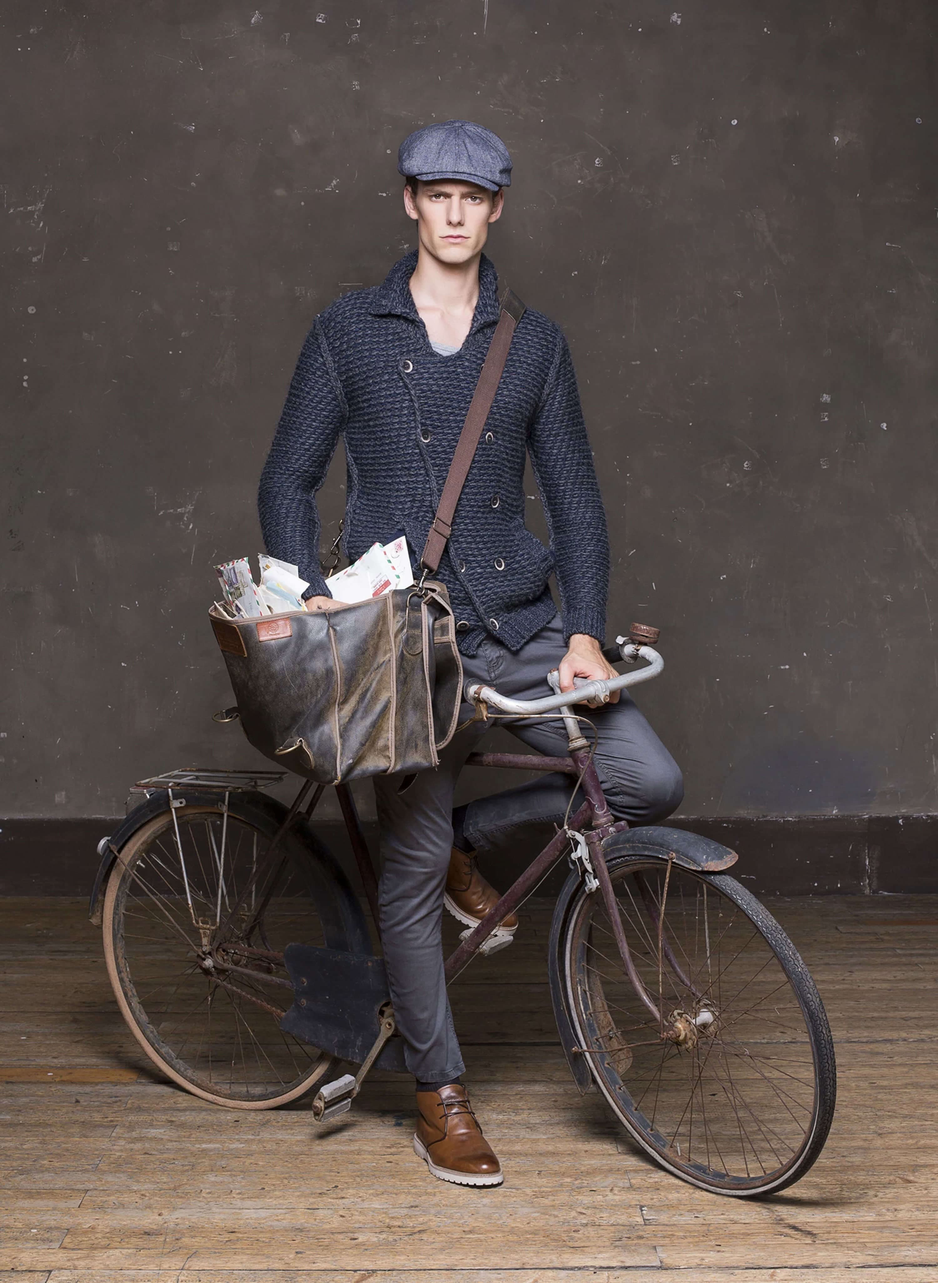 Full-length shot — the model as a postman: flat cap, chunky double-breasted knit cardigan, canvas satchel full of letters, leaning on an old bicycle