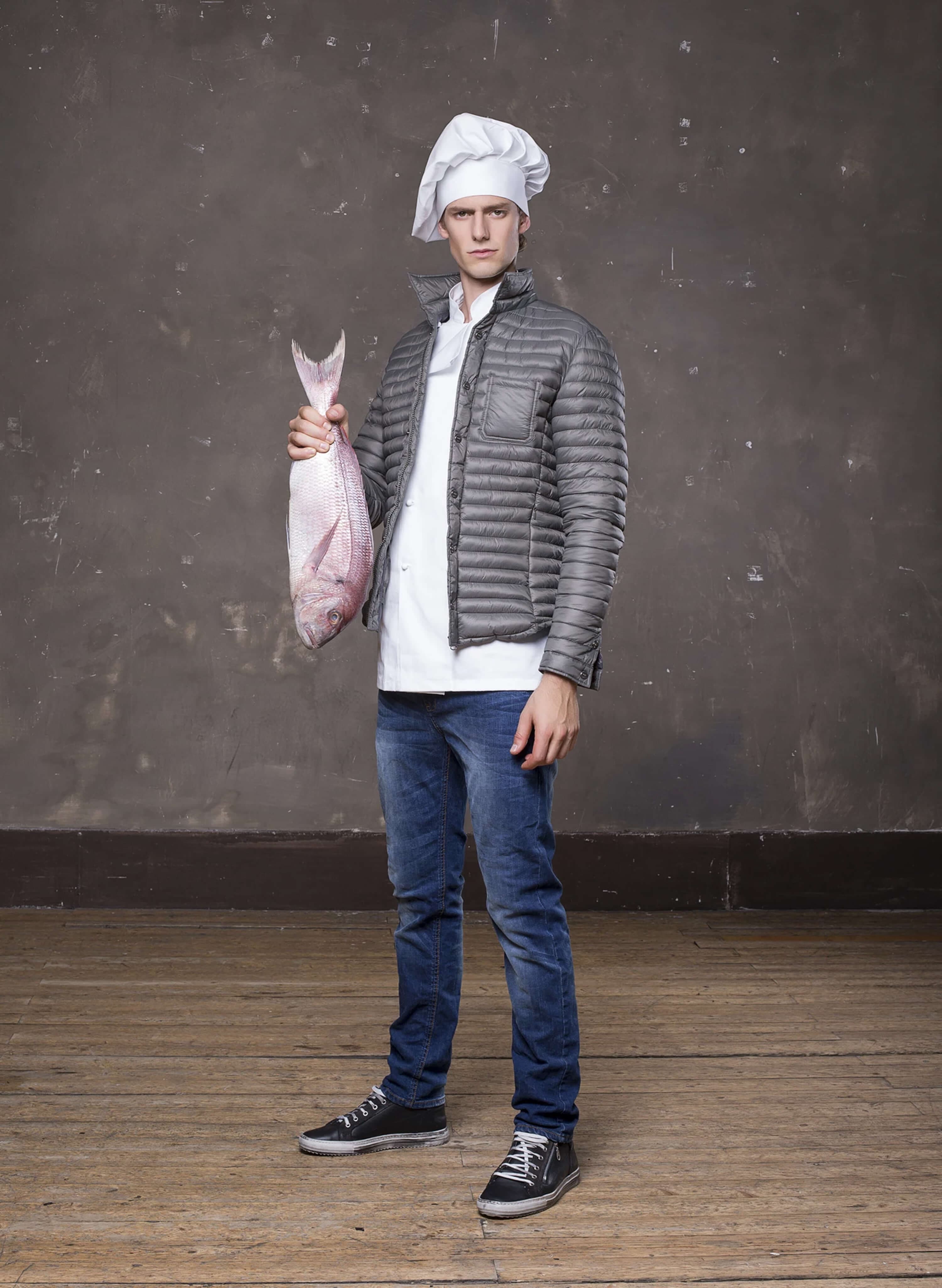 Full-length shot — the model as a chef: white toque and chef's jacket under a grey quilted down jacket, jeans and black plimsolls, holding a large raw fish by the tail