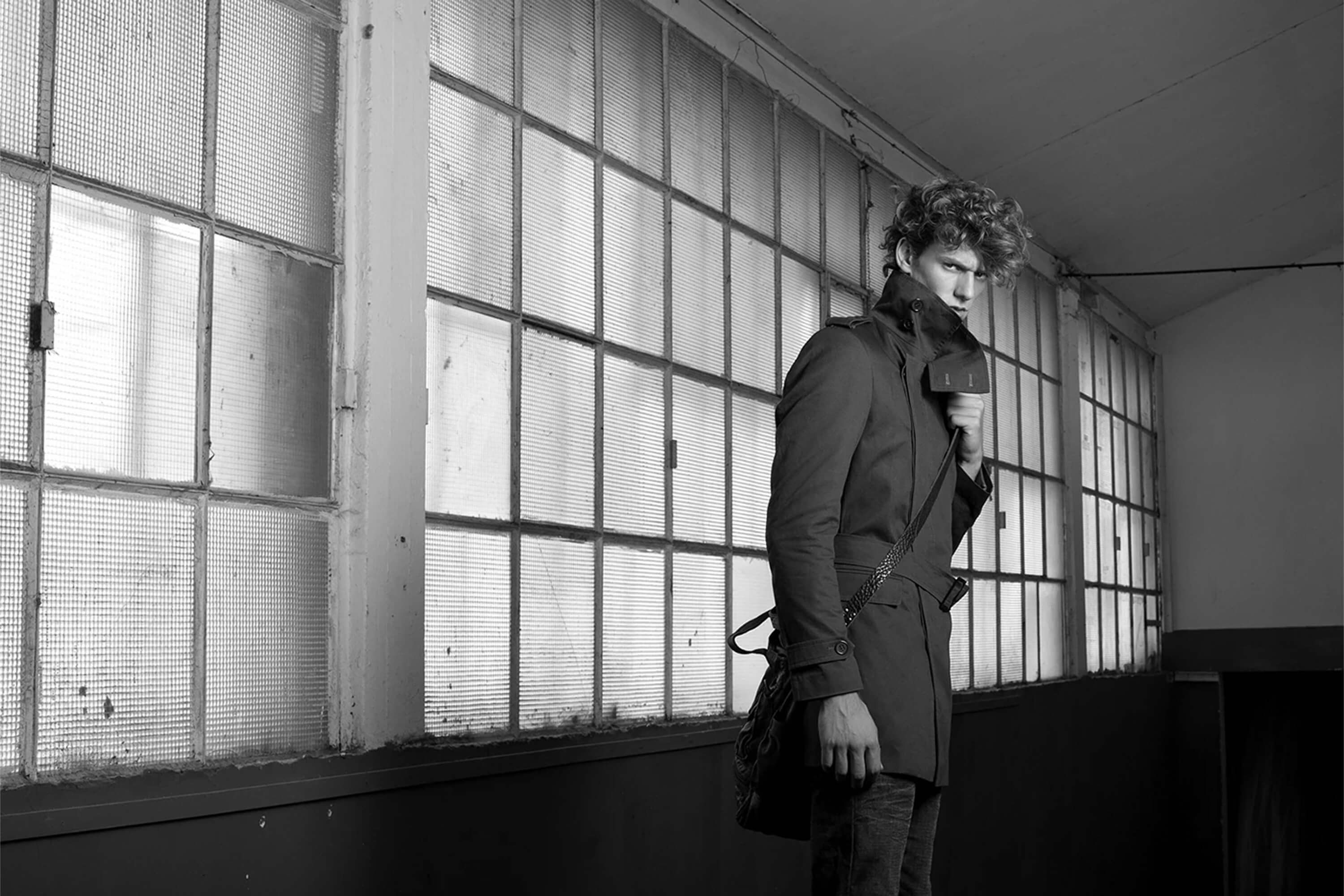 Black and white — the model in a belted trench coat with a shoulder bag, standing side-on along a wall of industrial glass-brick windows