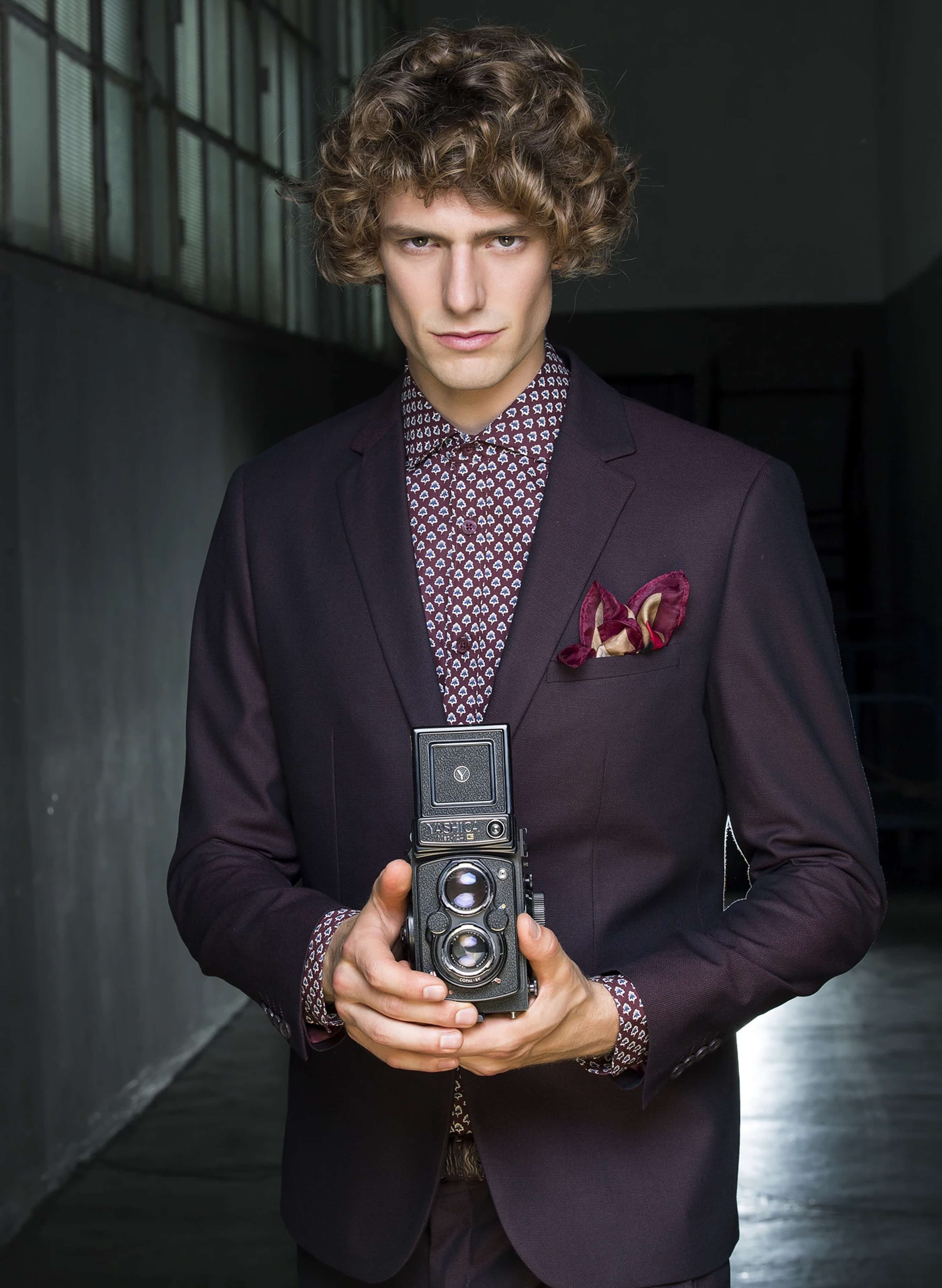 The model in a burgundy suit and patterned shirt with a silk pocket square, holding a vintage twin-lens Yashica camera at chest height
