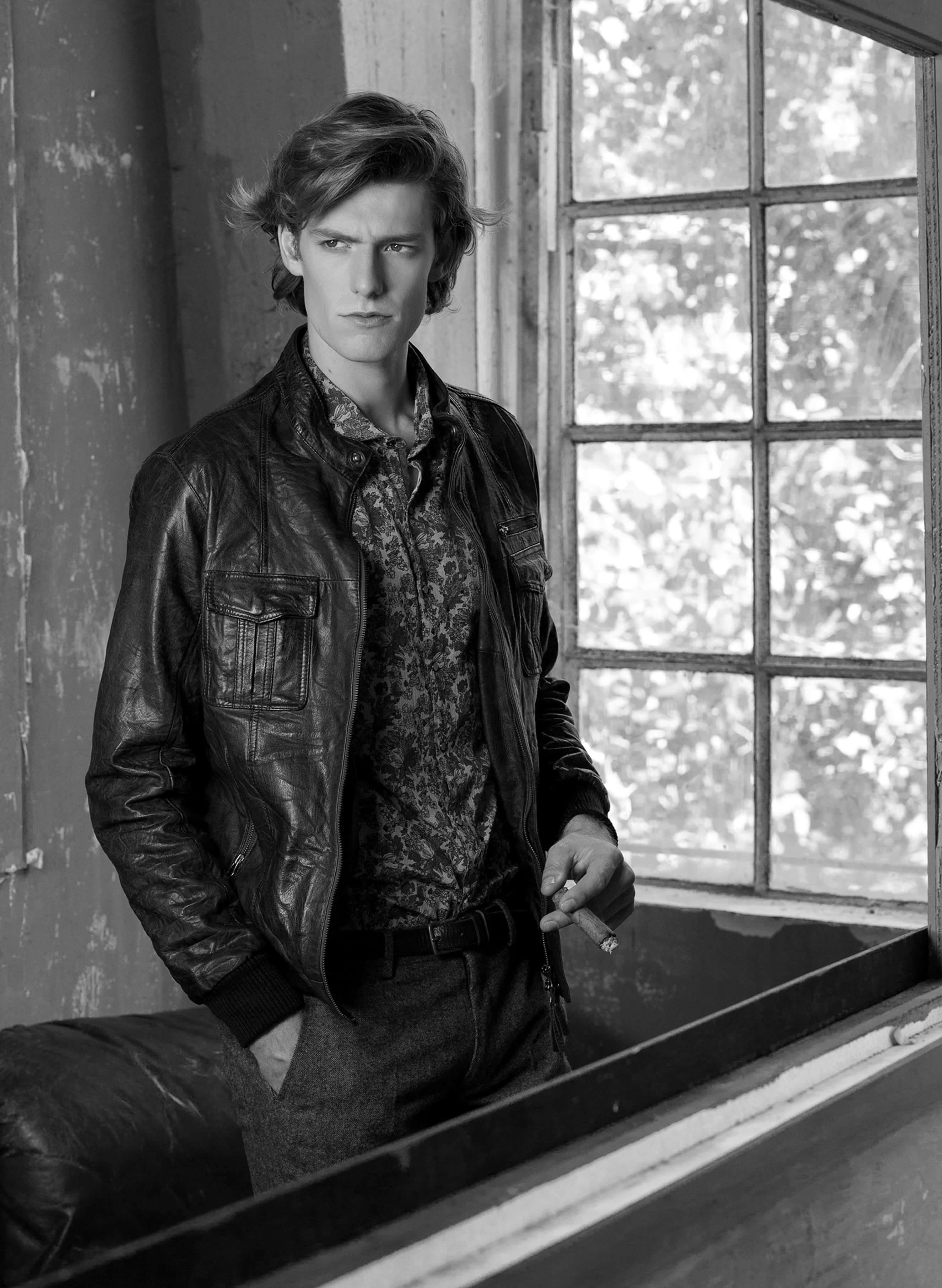 Black and white — the model in a leather bomber over a floral shirt, leaning against a window frame with a cigarette, the garden out of focus behind