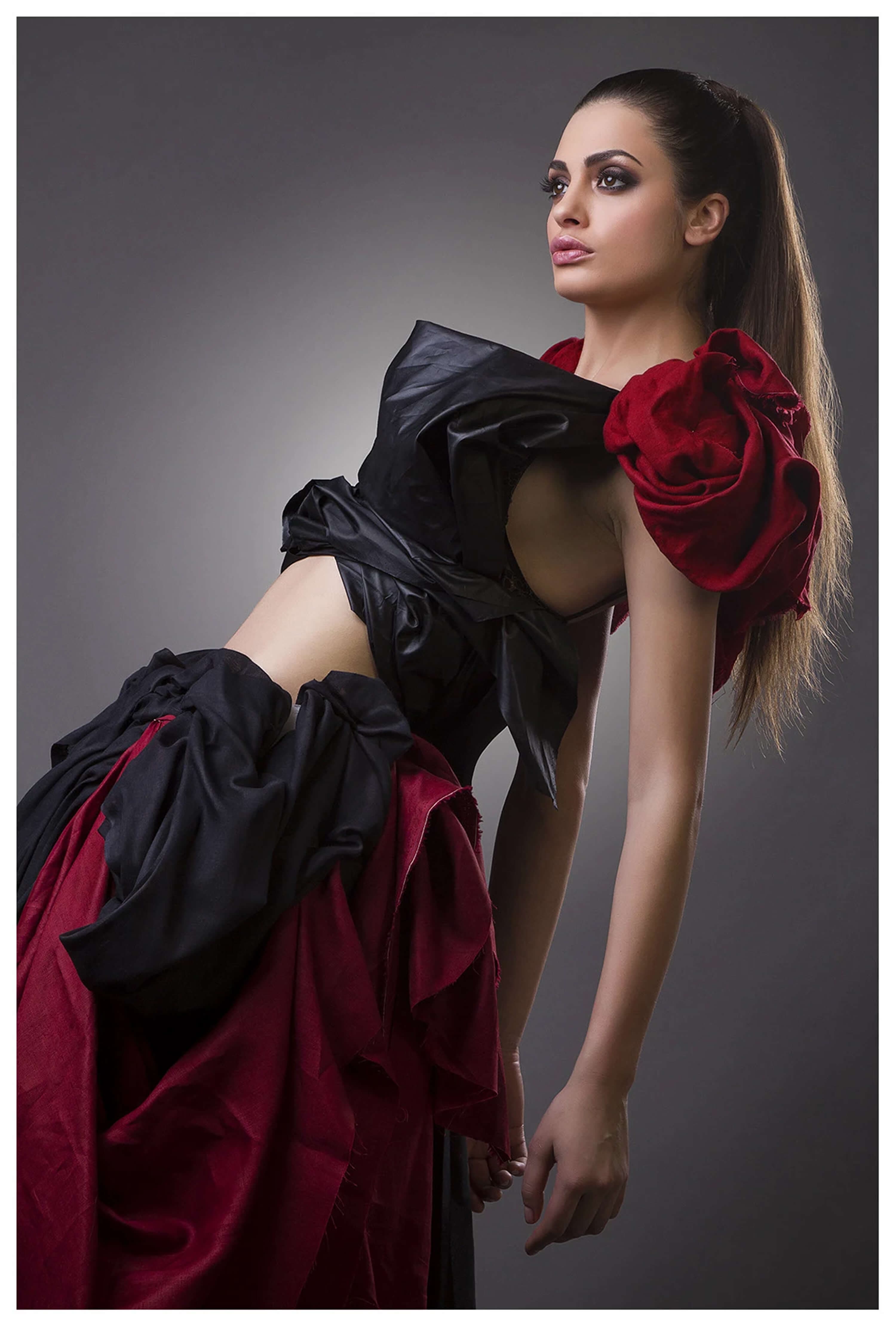 Leaning pose — black silk bodice with a deep red rolled shoulder, red and black draped skirt, the model turned away with her hair in a high ponytail
